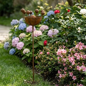 May include: A tall, rusty brown metal garden ornament with a bowl-shaped top and spherical accents. The ornament is set in a lush garden with vibrant pink, blue, and red flowers, and green foliage.
