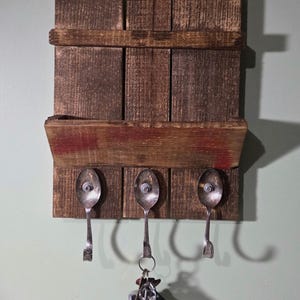 May include: A rustic wooden key holder with three repurposed spoon hooks. The wood is stained a dark brown with a red accent. Keys hang from the center spoon. A unique and functional home decor item.