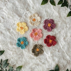 May include: A collection of seven crocheted flower appliqués arranged in a circular pattern. Each flower features five rounded petals in distinct colours including cream, yellow, pink, teal, dark red, olive green, and deep purple, all with a small yellow centre.