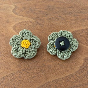 May include: Two five-petaled crochet flowers made of sage green yarn. The flower on the left features a bright yellow yarn center, while the flower on the right is centered with a smooth, round, black four-hole plastic button.