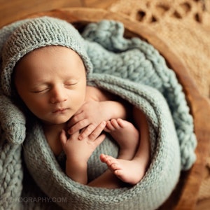 May include: A newborn baby wrapped in a soft, light blue knitted blanket and matching hat, nestled in a wooden bowl. The baby is sleeping peacefully, with tiny hands and feet visible. The background features a woven mat.