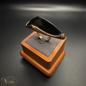 May include: A gold-toned ring featuring a large, smooth, black triangular stone set within a polished metal frame. The ring has a wide, curved design that tapers to a thin band, showcasing a sleek, reflective surface and a metallic gold finish.