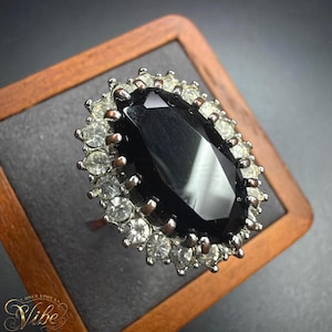 May include: A silver-toned ring featuring a large, oval-shaped, faceted black center stone surrounded by a halo of small, clear, round-cut white stones. The metal setting has a dark, oxidized finish. The text "ONCE UPON A VIBE" is visible in the corner.
