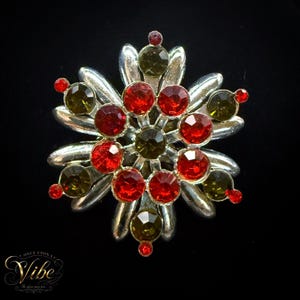 May include: A silver-toned brooch shaped like a star or snowflake, featuring a central dark olive-green crystal surrounded by a ring of bright red crystals, with additional olive-green and red crystals set along the radiating metallic arms.