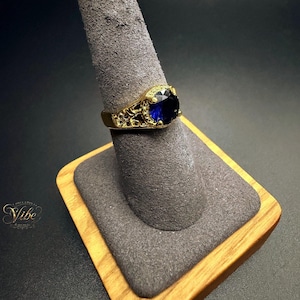 May include: A gold-toned ring featuring a large, round, faceted deep blue stone set in a decorative, openwork metal band with intricate filigree-style detailing along the sides, displayed on a gray velvet-textured conical ring stand.