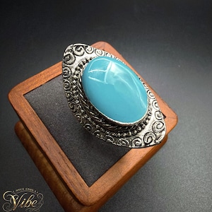 May include: A ring featuring a large, smooth, oval-shaped turquoise-colored stone set in a textured, antiqued silver-tone metal base. The metal setting displays intricate swirling patterns and a beaded border surrounding the central cabochon stone.