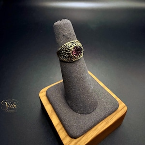 May include: A gold-toned ring featuring an intricate, openwork filigree band and a central, oval-shaped, deep purple gemstone. The metalwork displays a textured, detailed pattern surrounding the stone, which is set securely within the band.
