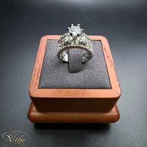 May include: A silver-toned ring featuring a prominent round-cut central stone flanked by smaller stones on each side. The band is encrusted with multiple tiny clear stones, creating a sparkling effect across the entire surface of the metal ring.