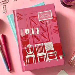 May include: A pink hardcover book featuring a graphic illustration of three chairs, a window with hanging laundry, and the text "Do more of what you love." The book is layered over a light blue notebook, surrounded by two pens, two pencils, and paper clips.