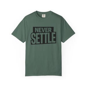 May include: A sage green short-sleeve t-shirt featuring the text NEVER SETTLE printed in bold black capital letters across the chest. The shirt has a crew neckline and a standard fit, with a small white brand label visible at the inner collar.