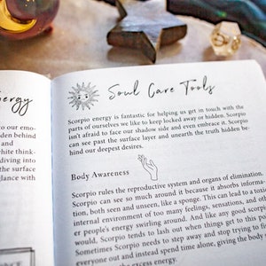 May include: An open book with the title "Soul Care Tools" and text about Scorpio energy and body awareness. The book is on a surface with decorative items, including a star and a moon-shaped object.