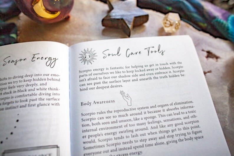 May include: An open book with the title "Soul Care Tools" and text about Scorpio energy and body awareness. The book is on a surface with decorative items, including a star and a moon-shaped object.