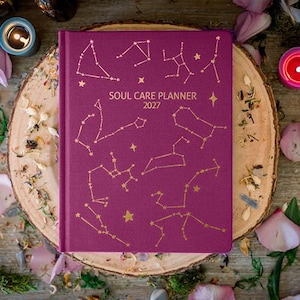 May include: A deep magenta hardcover planner with gold foil constellations and stars printed on the front cover. The text "SOUL CARE PLANNER 2027" is centered in gold lettering. The planner has a rectangular shape with a bound spine.