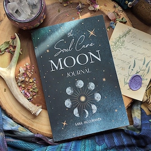 May include: A dark blue journal with a gold and white design. The cover features the text "The Soul Care Moon Journal" and a graphic of a moon with stars. The author's name, "Sara McCormick", is printed at the bottom of the cover.
