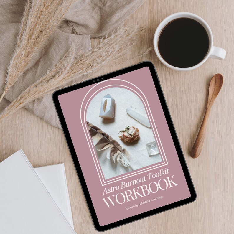 Puede incluir: Una tableta digital muestra la portada de un "Astro Burnout Toolkit WORKBOOK" con cristales y una pluma. Una taza de caf&eacute; y una cuchara de madera est&aacute;n sobre una superficie de madera clara. Tambi&eacute;n hay plantas secas y una pila de papel blanco.