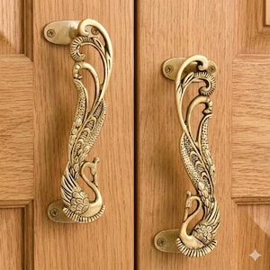 May include: Two ornate brass cabinet handles. Each handle features a swan design with intricate detailing. The handles are mounted on a light brown wooden cabinet door, adding a touch of elegance to the furniture.