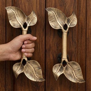 Peut inclure: Une paire de poignées de porte verticales en laiton avec une finition dorée métallique. Chaque poignée présente une barre cylindrique centrale flanquée d'éléments décoratifs en forme de feuille en haut et en bas, fixée par des plaques noires.