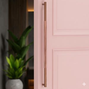 May include: A copper-colored door pull with a vertical design, mounted on a light pink door. The pull has a cylindrical shape with a darker copper accent at the top and bottom. The door has a panel design.