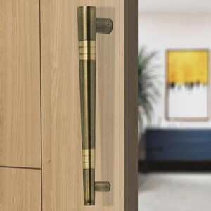 May include: A bronze-colored door pull with a cylindrical design, featuring gold-colored bands. The handle is mounted on a light-colored wooden door, with a glimpse of a room in the background.