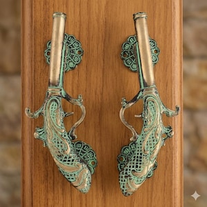 Puede incluir: Dos manijas de puerta de latón de estilo antiguo con forma de pistolas, montadas sobre una superficie de madera. Las manijas tienen detalles intrincados y un acabado de verdín, lo que aumenta su atractivo vintage. Montadas verticalmente.