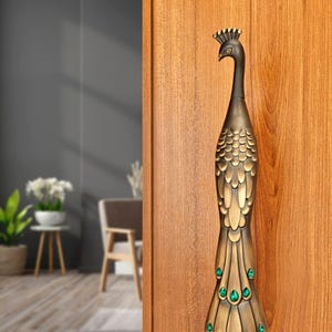 May include: A bronze-colored peacock-shaped door handle. The handle features a detailed design with a peacock's head, body, and tail. Green jewel-like accents are placed on the tail. The handle is mounted on a wooden door.