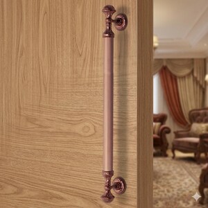 May include: A bronze-colored door pull with a vertical, ribbed design. The handle is mounted on a wooden door and features ornate, decorative end pieces. The door pull is a classic design element for interior doors.