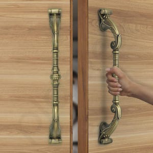 May include: Ornate bronze door handles with a detailed, vintage design. The handles feature a vertical, elongated shape with decorative elements and are mounted on a light brown wooden door. One handle is being held by a person.