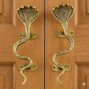 Puede incluir: Dos manijas de puerta de latón con forma de cobra. Cada manija presenta una cabeza de cobra detallada y un cuerpo sinuoso, montadas sobre una placa rectangular. Están fijadas a una puerta de madera, mostrando un diseño decorativo.