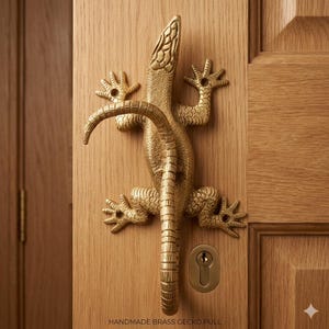Puede incluir: Un tirador de puerta de gecko de latón hecho a mano, montado en una puerta de madera. El gecko dorado presenta escamas detalladas y un asa curvada. El texto "HANDMADE BRASS GECKO PULL" está en la parte inferior.