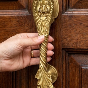 Puede incluir: Un tirador de puerta de latón pulido con una cabeza de león detallada en la parte superior, un eje central con diseño de cuerda trenzada y una base decorativa en forma de hoja. El herraje está montado verticalmente en una puerta de madera.