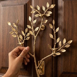 May include: A gold-toned metal wall hook shaped like a branching tree with three small birds perched on the limbs. The structure features textured bark details and numerous leaves, with two circular mounting plates attached to the wooden door surface.
