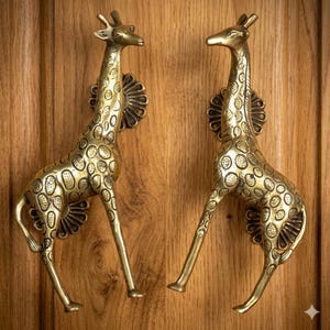 Puede incluir: Dos tiradores de puerta de latón con forma de jirafa montados en una puerta de madera. Las jirafas tienen manchas detalladas y están de pie. Los tiradores son decorativos y añaden un toque único a cualquier puerta.