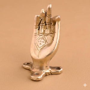 Peut inclure: Sculpture en forme de main en laiton, avec des détails fins et un motif en losange sur la paume. La main est en position de bénédiction, montée sur une base avec trois trous. L'objet présente un aspect vintage et une finition dorée.