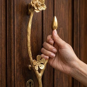 Puede incluir: Un tirador de puerta de metal dorado con un tallo texturizado en forma de rama, una flor grande en la parte superior y una flor más pequeña de cinco pétalos cerca de la base, montado en una puerta de madera marrón oscuro con una cerradura.