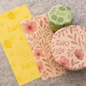 May include: An overhead shot of several reusable food wraps. One is yellow with bee and honeycomb graphics, and the text "ZaiQ". Two wraps have floral patterns, and two round containers are also decorated with floral patterns and the text "ZaiQ".