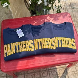 May include: Three navy blue crewneck sweatshirts featuring the word PANTHERS in large, yellow and white gingham-patterned block letters across the chest. The leftmost sweatshirt includes a small white rectangular tag attached to the collar.