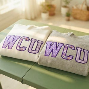 May include: A white hoodie and a gray hoodie, each featuring the letters WCU in large, purple, patterned applique. The letters are decorated with a mix of polka dot, floral, and gingham prints, with a thick purple border around each letter.