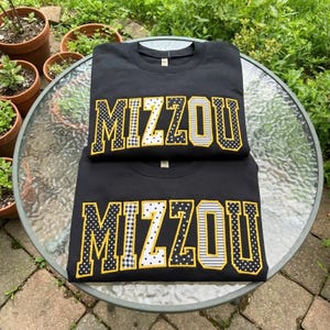 May include: Two black crewneck sweatshirts stacked on top of each other. Each sweatshirt features the word MIZZOU in large block letters across the chest. The letters are outlined in yellow and filled with various patterns including polka dots, stripes, and gingham.