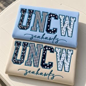 May include: Two folded sweatshirts, one light blue and one beige, featuring the text UNCW in large block letters with varied patterns including floral, gingham, and stars, and the word seahawks embroidered in cursive script below the letters.