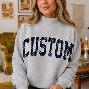 May include: A light gray long-sleeve sweatshirt with a mock neck collar. The front features the word CUSTOM in large, dark navy blue block letters with a white polka dot pattern and a thick white outline, centered across the chest.