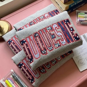 May include: A light gray sweatshirt featuring the word DUQUESNE in large, block-style letters. Each letter is outlined in red thread and filled with a variety of patterned fabrics, including floral, gingham, striped, and star-print designs.