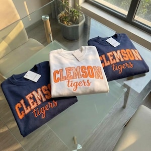 May include: Three crewneck sweatshirts in navy blue and white, each featuring the text CLEMSON tigers in orange block and script lettering. The orange letters have a subtle checkered pattern. Each garment includes a white paper tag at the collar.