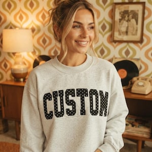 May include: A light gray crewneck sweatshirt featuring the word CUSTOM printed in large, bold, black block letters with a white polka dot pattern. The garment has long sleeves, a ribbed neckline, and a relaxed fit, providing a classic look.