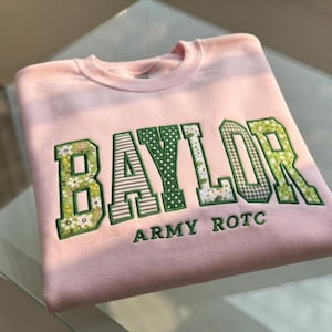 May include: A light pink crewneck sweatshirt featuring the word BAYLOR in large, block-style letters with various green and white floral, striped, and polka-dot patterns. Below the main text, the words ARMY ROTC are embroidered in green thread.