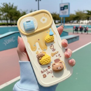 May include: A cream-colored phone case with a yellow and brown bear theme. The case features 3D bear and honeycomb designs, with the words "Nice Day" and "Bear" printed on the case. A small blue and white milk carton design is also present.