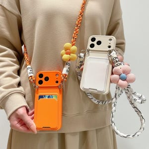 May include: Two mobile phone cases with integrated card slots and crossbody straps. One case is bright orange with an orange braided strap and yellow flower charm. The other is cream with a white braided strap and a pink flower charm.