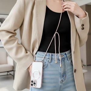 May include: A clear smartphone case with a metallic pink circular magnetic ring on the back, attached to a long, adjustable pink cord strap with metallic end caps, worn as a crossbody accessory over a black top, blue jeans, and a beige blazer.