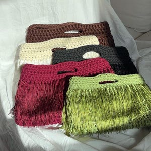 Könnte beinhalten: Eine Kollektion von fünf rechteckigen Häkeltaschen mit integrierten Griffen und langen, fließenden Fransen. Die Taschen sind in verschiedenen Unifarben erhältlich, darunter Dunkelbraun, Creme, Anthrazit, tiefes Burgunderrot und Limettengrün.