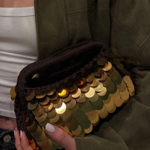 May include: A dark brown crochet clutch bag covered in overlapping, reflective gold circular sequins. The bag features a thick, textured dark brown crocheted rim and a structured, rounded shape, showcasing a metallic finish that catches the light.