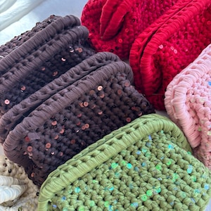 May include: A collection of rectangular crochet clutch bags in various colors including red, dark brown, light pink, olive green, and cream. Each bag features a thick, textured knit pattern embellished with small, shimmering sequins.